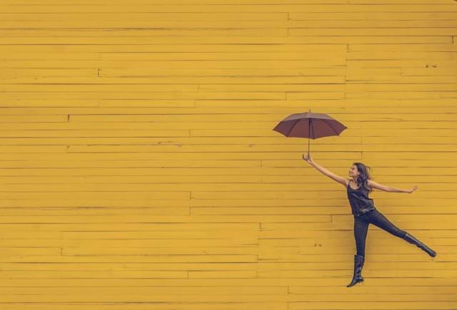 Image of a woman with an umbrella floating upwards against a yellow wall 